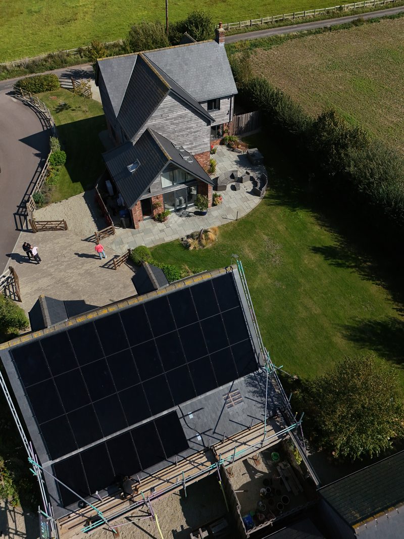 Aerial view of a steep slate roof densely fitted with black solar panels on a rural chalet-style home, scaffolding still in place