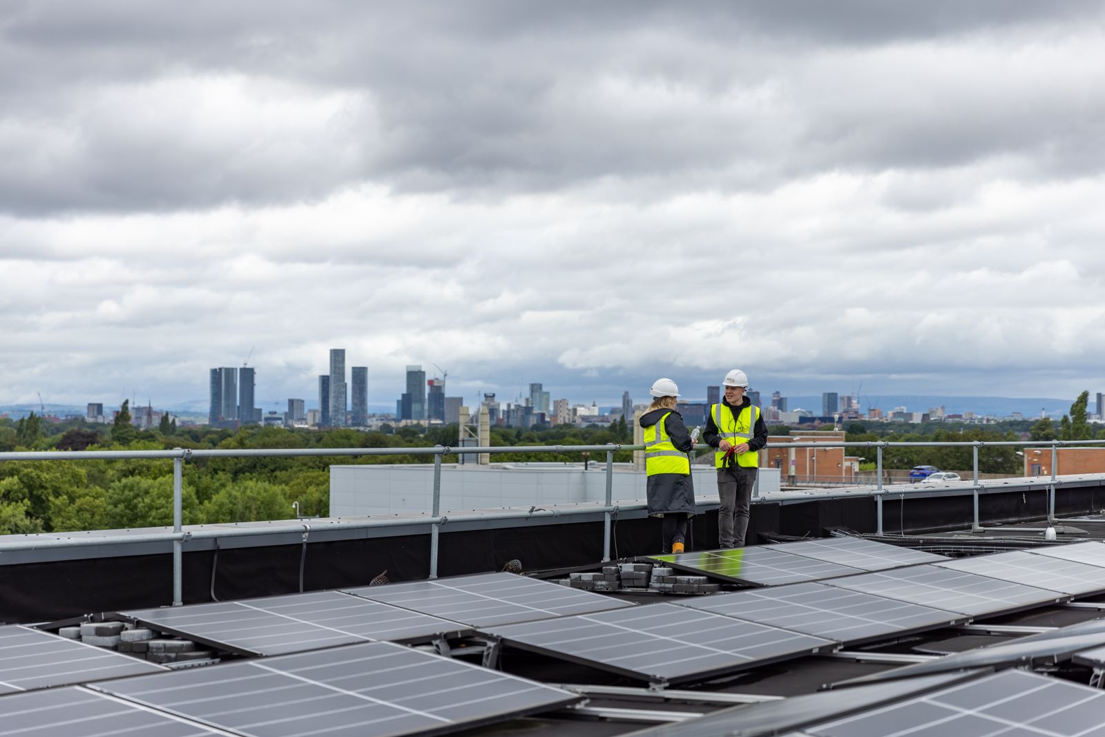 Two installation team members in hard hats and hi-vis on a solar roof, Manchester skyline behind